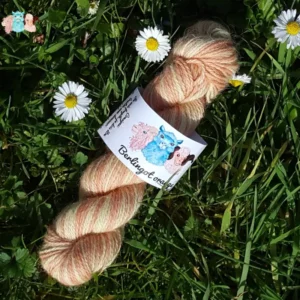 Echeveau de laine alpaga, teintée naturellement en nuances orangé et jaune tendre posée sur de l'herbe verte avec des pâquerettes. Laine artisanale, teinture végétale faite à la main en France