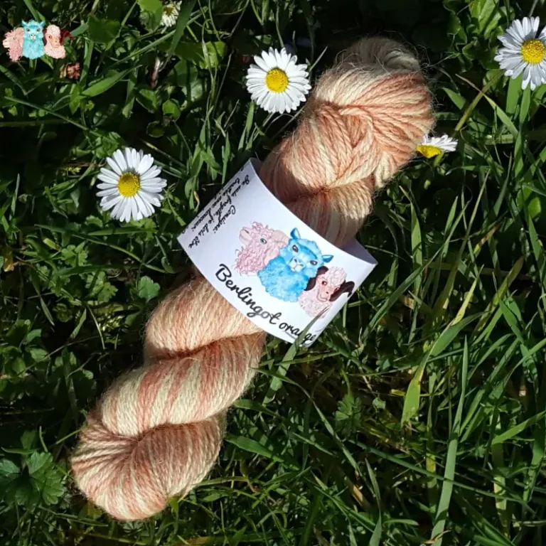 Echeveau de laine alpaga, teintée naturellement en nuances orangé et jaune tendre posée sur de l'herbe verte avec des pâquerettes. Laine artisanale, teinture végétale faite à la main en France