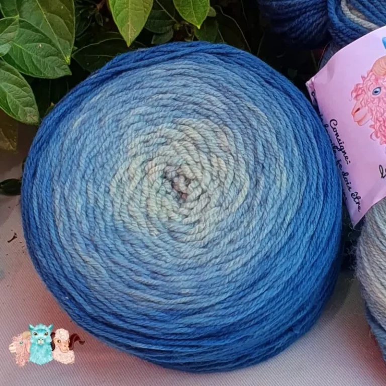 Détail d'une pelote de laine de coloris du bleu foncé au bleu clair devant des feuilles vertes et posé sur un fond gris.