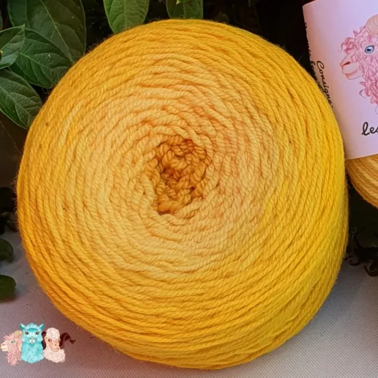 Détail d'une pelote de laine de coloris du jaune foncé au centre au jaune intense devant des feuilles vertes et posé sur un fond gris.