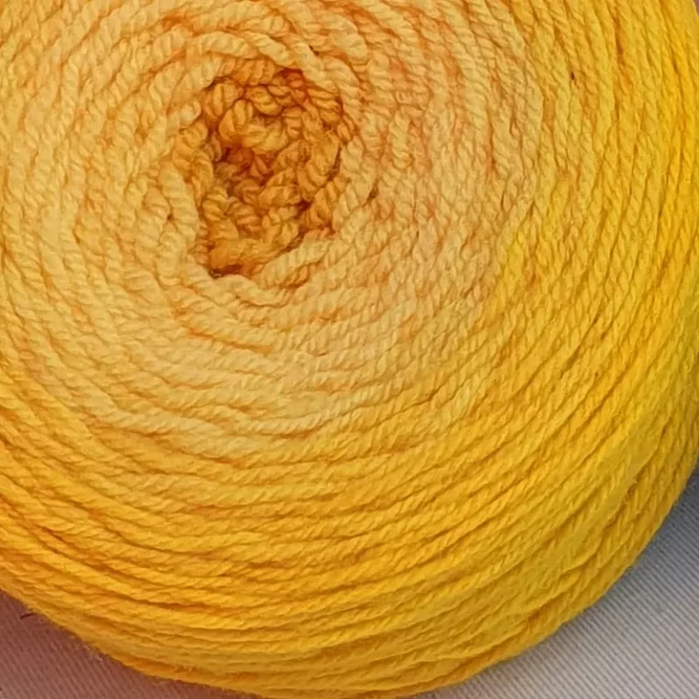 Gros plan d'une pelote de laine de coloris du jaune foncé au centre au jaune intense devant des feuilles vertes et posé sur un fond gris.