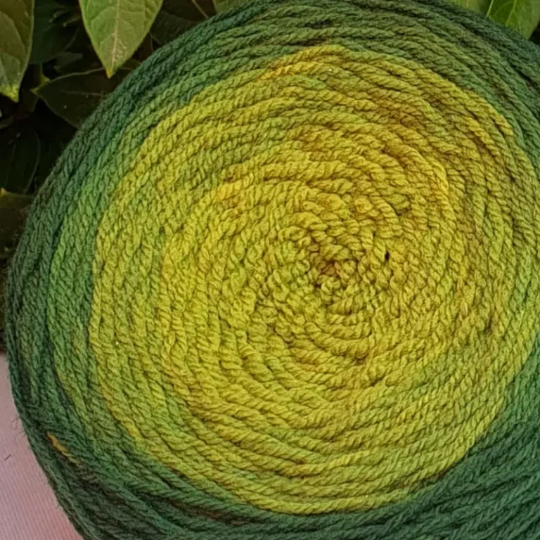 Gros plan d'une pelote de laine de coloris du vert sombre au vert clair devant des feuilles vertes et posé sur un fond gris.