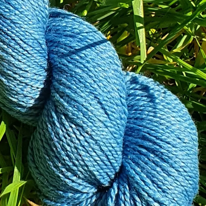 Gros plan d'un écheveau de laine mérinos soie teinté en bleu sur un fond d'herbe verte.