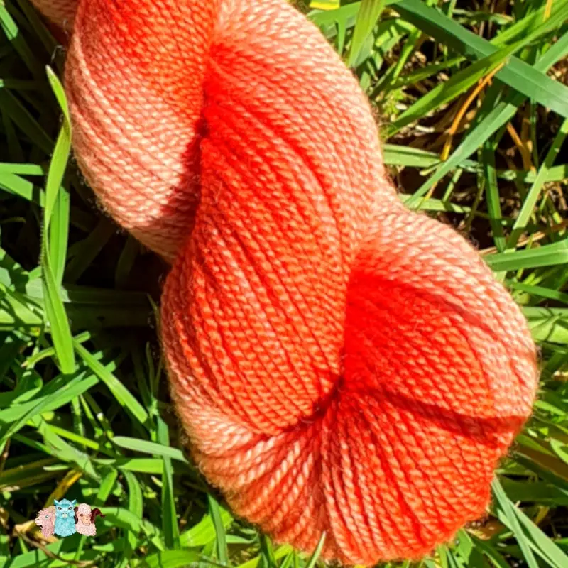 Gros plan d'un écheveau de laine mérinos soie teinté en orange nuancé sur un fond d'herbe verte.