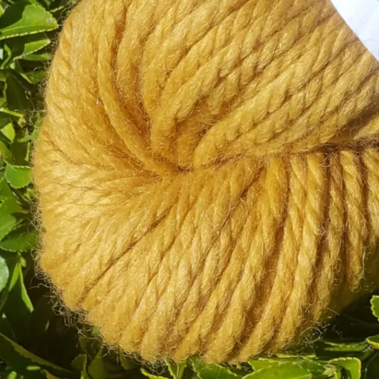 Gros plan d'un écheveau de laine mérinos alpaga teinté en jaune doré sur un fond de feuilles vertes.