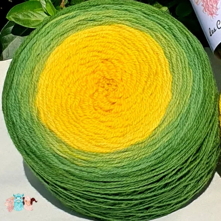 Détail d'une pelote de laine de coloris du vert sombre au jaune intense devant des feuilles vertes et posé sur un fond gris