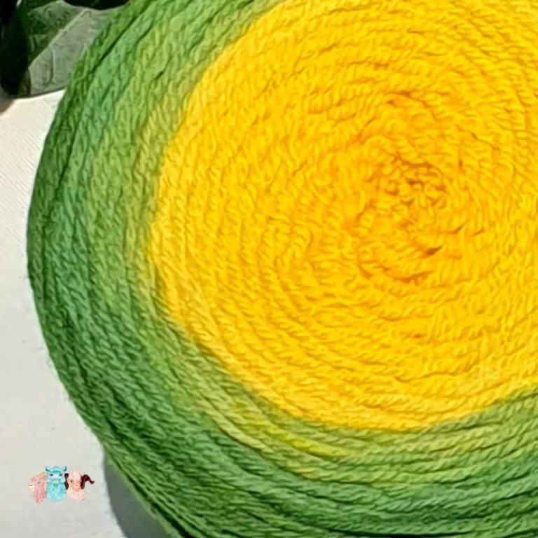 Gros plan d'une pelote de laine de coloris du vert sombre au jaune intense devant des feuilles vertes et posé sur un fond gris