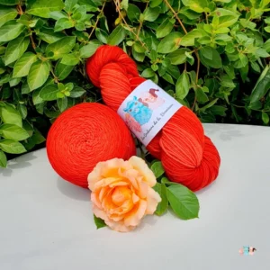 Echeveau et pelote de laine de coloris de l'orange foncé à l'orange rouge clair devant des feuilles vertes et posé sur un fond gris avec une rose.