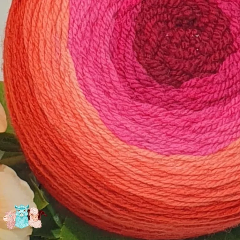 Gros plan pelote cake de laine de coloris de l'orange foncé au rouge bordeaux devant des feuilles vertes et posé sur un fond gris avec une rose.