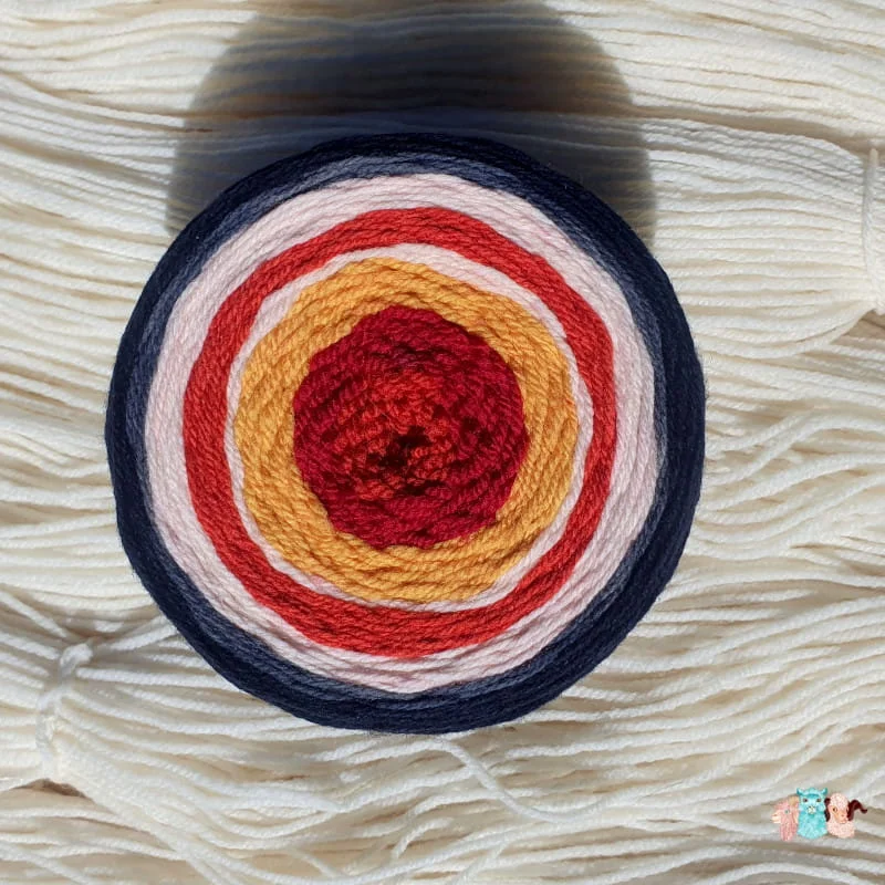 Détail d'une pelote cake de laine de coloris orange, rouge, jaune , écru, orange, écru et noir sur un fond de fil de laine écrue.