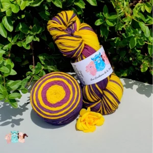 Echeveau et cake de laine de coloris violet et jaune devant des feuilles vertes et posé sur un fond gris avec une rose.
