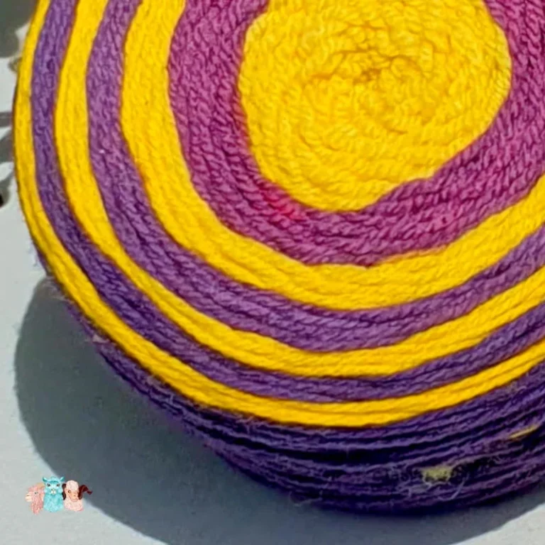 gros plan d'un cake de laine de coloris violet et jaune devant des feuilles vertes et posé sur un fond gris avec une rose.