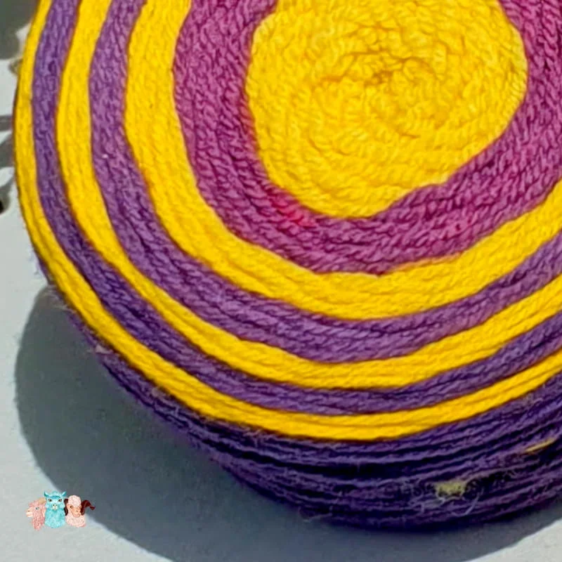 gros plan d'un cake de laine de coloris violet et jaune devant des feuilles vertes et posé sur un fond gris avec une rose.